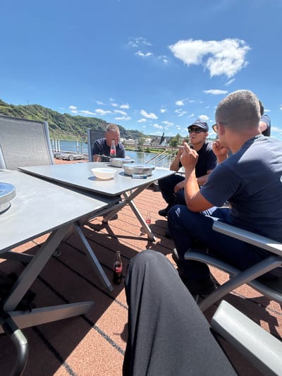 The media file depicts an outdoor scene with four individuals seated around a table, seemingly engaged in a meal or social gathering. The setting appears to be a deck or patio overlooking a body of water, with a lush, tree-covered hillside in the background. The sky is a vibrant blue, dotted with fluffy white clouds, suggesting a clear, pleasant day. Sunlight casts distinct shadows, indicating it is likely midday.
The individuals are casually dressed. Two are seen from behind, one with a dark blue t-shirt and navy trousers, the other in a similar dark blue shirt and dark pants. A third person, facing forward and partially obscured, wears a dark t-shirt and a baseball cap. The fourth person, seen in profile, is wearing a dark t-shirt and has short, curly hair. One individual is holding a utensil and appears to be eating or interacting with food on their plate. Another person is drinking from a bottle of Coca-Cola, which is visible on the table.
Objects on the table include plates, bowls, and a bottle of Coca-Cola. The table itself is a light gray, rectangular surface, supported by metal legs. The chairs are also made of metal with gray fabric seats. The ground beneath the table and chairs is a textured reddish-brown material, possibly artificial turf or a similar outdoor flooring.
A prominent detail is a bottle of Coca-Cola with its distinctive red label, clearly visible on the table and on the ground. The surrounding environment, with the river or canal, the hillside, and architecture visible in the distance, hints at a European setting. Given the context of Piesport, Germany, this could be a riverside restaurant, a private terrace, or a boat deck. The overall mood seems relaxed and convivial.