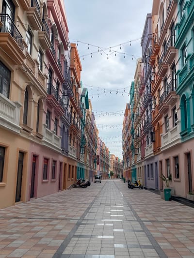 The image shows a street in Nghĩa Trụ Commune, Vietnam, lined with colorful buildings. The buildings are painted in pastel shades of pink, peach, lavender, and teal, with architectural details like balconies, arched windows, and decorative cornices. String lights are hung overhead, illuminating the walkway. The street is paved with patterned tiles, and there are a few motorcycles parked on the side. A white van is visible in the distance. The sky is overcast.