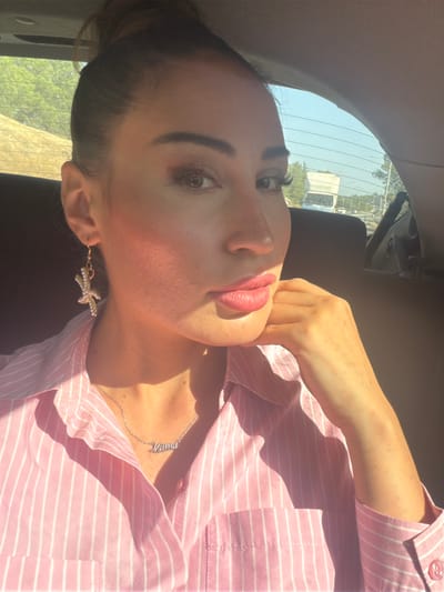 This is a slightly overexposed, close-up, portrait-style photograph of a woman. The image is oriented vertically, and the woman is looking towards the viewer with her head tilted. She has dark hair pulled back, and she is wearing makeup, including dark eyeliner and lipstick. She has a delicate necklace with a silver pendant that appears to spell "Mama" and a starfish-shaped earring. She is wearing a light pink and white striped button-up shirt. Her right arm is bent, with her hand resting near her chin. Sunlight is prominently featured, casting shadows across her face and highlighting her skin.

The background suggests an indoor setting, possibly a car, with a window showing a sunny outdoor scene. Trees and a building are visible through the window, indicating a daytime setting with clear weather. The overall mood is relaxed and casual. The sunlight suggests it is a bright day, possibly late morning or afternoon. The provided location context is Aradippou, Cyprus, which aligns with the bright, sunny weather depicted.