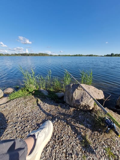 The image shows a lakeside scene in Gargždai, Lithuania. A person's leg and foot, wearing a white sneaker and grey pants, are visible in the foreground, suggesting they are sitting on the shore. A fishing rod with a blue and green reel lies on the ground next to a large rock and tall grass. The lake stretches out in the background, with a treeline and a few buildings visible on the opposite shore under a clear blue sky with scattered white clouds. The ground is covered with pebbles and some vegetation.