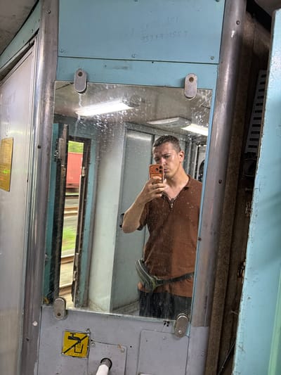 In Saphale, India, a man takes a mirror selfie inside what appears to be a train compartment. The man, wearing a brown polo shirt and dark pants, holds an orange smartphone to capture his reflection. He has short hair and a serious expression. The compartment features a large, grimy mirror that reflects him and the interior of the train. Visible in the reflection are fluorescent lights, metal fixtures, and a sliver of the outside world, showing greenery and train tracks through an open door. The overall environment is utilitarian, with faded blue and gray paint, metallic poles, and worn surfaces, indicating the age and use of the train car.