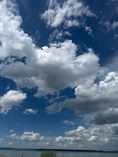 This media file depicts a wide-angle view of a vast blue sky filled with a variety of clouds, ranging from bright white cumulus clouds to darker, more ominous storm clouds. The clouds are dynamic and textured, with some appearing wispy and scattered while others are dense and voluminous.

The bottom portion of the image reveals a landscape that appears to be a body of water, possibly a lake or river, meeting a distant shoreline. The shoreline is characterized by a thin strip of trees or vegetation. The overall impression is one of a natural, open environment, likely a rural or undeveloped area.

The lighting suggests it is daytime, with bright sunlight illuminating the clouds and water. The presence of varied cloud formations hints at dynamic weather conditions, possibly a mix of sunshine and approaching rain.

The provided location context, Володарский ауылдық округі, Kazakhstan, suggests the scene is set in a rural area of Kazakhstan, characterized by open skies and bodies of water. The image evokes a sense of spaciousness and the raw beauty of nature. There is no visible text in the image.