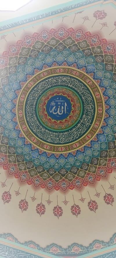An overhead view captures a vibrantly decorated domed ceiling or a large circular wall panel, likely within a mosque in Mns Asan, Indonesia. The central motif is a circular medallion featuring the Arabic word "الله" (Allah) in white Thuluth script on a light blue background, with "محمد" (Muhammad) and "منون" (Manoon/Mnon) inscribed below it. This central element is encircled by a series of concentric rings. The innermost rings display intricate geometric patterns in red and green, followed by a dark teal ring containing white Arabic calligraphy, potentially the Asmaul Husna (99 Names of Allah), and then a gold-yellow ring with red and blue decorative elements.
Beyond these rings, the design expands radially outward with numerous fan-shaped or scalloped panels. Each panel contains white Arabic calligraphy on backgrounds of varying hues, including blues, greens, browns, and muted reds, forming a repetitive, intricate pattern. These individual motifs are arranged in an expanding arc across the visible surface. The outermost visible sections feature lighter, more sparsely arranged decorative elements and pendant-like shapes in red, pink, and teal on an off-white background, leading to a light blue border with faint white script along the top edge of the frame. No individuals, furniture, or other objects are visible.