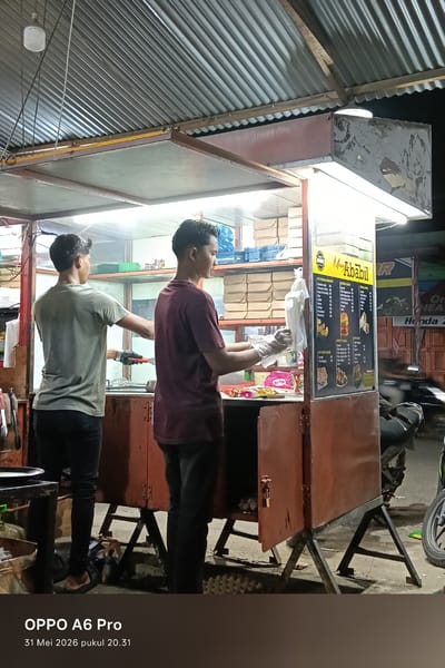 The image captures two men working at a street food stall in Sangso, Indonesia. The stall, named "Menu Ababil," displays a menu board and shelves stocked with food containers. One man, facing away, is wearing a light gray shirt and black pants, operating with black gloves. The other man, wearing a burgundy shirt and black pants, faces the stall and is wearing gloves. The structure is covered by a corrugated metal roof, and a motorcycle is parked nearby. The image is taken at night, under bright artificial lighting, with the text "OPPO A6 Pro" and the date "31 Mei 2026 pukul 20.31" at the bottom, suggesting the photo's origin or a timestamp.