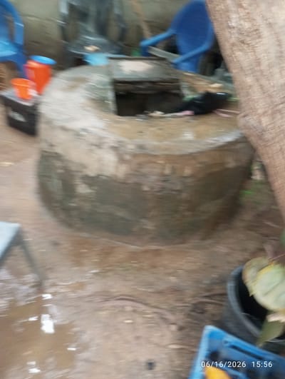 The image shows an outdoor scene with a well in the foreground. The well is circular and appears to be made of stone or concrete, with some vegetation growing around its rim. There are several objects scattered around the well, including plastic buckets, a blue chair, and what appears to be a tree trunk. The ground is wet and muddy, suggesting recent rain. In the background, there are some blurred structures that could be buildings or other outdoor features. The overall impression is that of a somewhat unkempt, rural or semi-rural environment. The timestamp at the bottom right of the image indicates the date and time as 06/16/2026 15:56. The location is identified as Dokan Tofa, Nigeria.