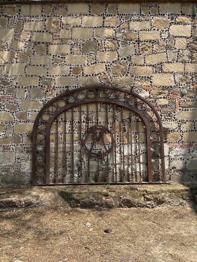 The image captures a weathered stone wall in Tlalnepantla, Mexico. The wall's construction consists of rectangular stones arranged in a grid pattern with smaller dark stones filling the gaps. An ornate, rusted metal gate is affixed to the wall, its design featuring a series of circles along the top arch and horizontal bars in the main section. A circular emblem is located in the center of the gate. To the right of the wall is a section of rough, earthen terrain covered in dry vegetation. The lighting is bright, casting shadows that accentuate the textures of the wall and the gate.