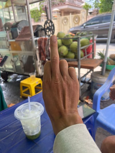 A person's hand, displaying a middle finger, is prominently featured in the foreground of the image. The hand appears to be resting on a blue plastic table. To the left of the hand is a clear plastic cup with a white lid and a straw, containing a green liquid with ice. In the background, there is a street scene with a cart laden with coconuts. The environment suggests an outdoor setting, possibly a market or roadside vendor. The specific location is identified as Binh Thuan Ward, Vietnam.