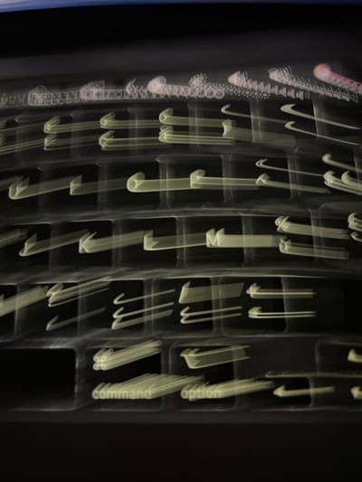 The image captures a backlit computer keyboard, likely a MacBook, rendered with significant horizontal motion blur. The scene is dark, suggesting low light conditions, possibly indoors in Buxerolles, France.
The primary subject is a QWERTY keyboard, its keys outlined and characters illuminated in a pale yellow-green. Due to the motion blur, individual keys appear as elongated horizontal streaks. Identifiable keys include "command" and "option" on the bottom row, and partially visible letters such as "M" and "K" in the middle rows. Above the main keyboard section, at the top of the frame, are multiple highly blurred, thin white and reddish light streaks. Some white streaks in the upper-left corner exhibit patterns resembling repetitive structures or interconnected shapes, possibly originating from a distant display or complex light source, also affected by the motion blur. No individuals are present. The overall environment appears to be an interior setting, devoid of specific discernible decor beyond the light sources and the keyboard.