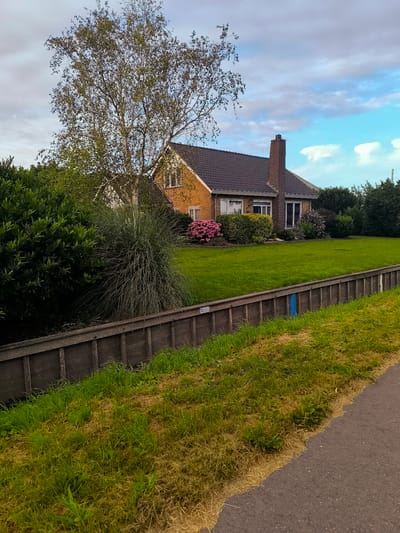 This image depicts a residential property with a brick house and a well-maintained lawn, situated in the city of Wervershoof, Netherlands. The house, constructed of orange brick with a dark tiled roof, features prominent windows and a brick chimney. It is surrounded by lush green grass, manicured hedges, and flowering bushes, including vibrant pink blooms. A tall, slender tree with sparse foliage stands to the left of the house. In the foreground, a wooden fence-like structure borders a grassy area, beyond which a paved path is visible. The sky above is a mix of blue and overcast clouds. The scene appears serene and domestic, with no people or animals present, and no discernible activity taking place. The overall impression is one of a tranquil, suburban setting.