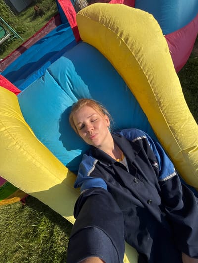 A woman with fair skin and reddish-blonde hair is lying on her back on a brightly colored inflatable structure, likely a bouncy castle, which is situated on grass outdoors. Her eyes are closed, and her lips are pursed, suggesting she is relaxing or sleeping. She is wearing a dark blue jacket with blue and white trim over a lighter colored shirt. The inflatable structure is composed of large, curved sections in yellow, blue, and red. Sunlight casts a shadow of the woman's profile onto the blue section of the inflatable. The background shows more green grass and possibly some outdoor equipment. The scene appears to be in a residential or recreational outdoor area.