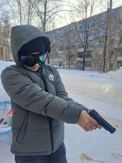 An outdoor scene in Bolshaya Elhovka, Russia, depicts a young individual standing on a snow-covered path. The person is clad in a dark green hooded puffer jacket featuring a visible "Kappa" logo on the left chest, a dark beanie, and dark trousers. Mirrored, rainbow-tinted goggles cover the upper face. The individual holds a black object resembling a semi-automatic pistol in a two-handed grip, with arms extended forward. The background displays a multi-story, light-colored apartment building with numerous windows. Bare deciduous trees are scattered throughout the snow-covered landscape. The ground consists of white snow, with visible patches of exposed brown earth or dirt on the path. A substantial snowdrift is present in the right foreground.
