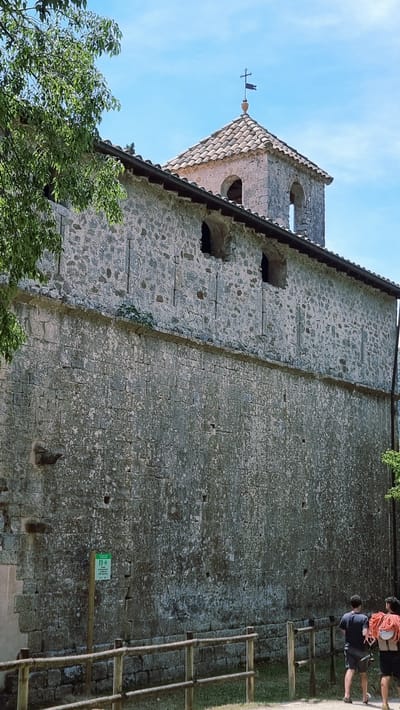 The image shows a stone building with a tiled roof and a bell tower topped with a cross. The building has a rustic appearance, with rough-hewn stone blocks and small, rectangular windows. In the foreground, a wooden fence runs along a path, and two people are visible walking away from the camera. One person is carrying a large coil of red rope over their shoulder. A green sign with white text and an arrow pointing right is mounted on a wooden post to the left of the fence. The sky is blue and clear, with some green foliage visible on the left side of the image. The scene appears to be outdoors, possibly in a rural or historical setting.