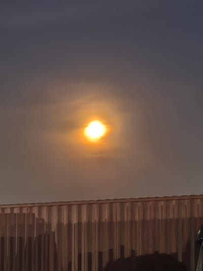 The image shows a sun or moon obscured by clouds, appearing as a bright, slightly blurred orange orb in the upper center of the frame. Below the sun/moon, a section of a corrugated metal fence or wall is visible. The sky is a gradient of grey and orange, suggesting twilight or dawn. The scene is likely located in an outdoor environment. I can not confirm it is Alcàsser, Spain.