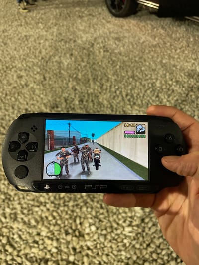 A hand holds a black PlayStation Portable (PSP) displaying the game "Grand Theft Auto: Vice City Stories." The game's screen shows the player character leading a group of soldiers on motorcycles. The PSP's interface elements, like time and money, are visible. The background features a patterned beige carpet. A portion of a black toy car is visible on the left side of the image.