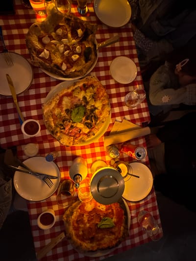The scene is an overhead view of a restaurant table located in Naples, United States of America. The table is draped with a red and white checkered tablecloth. Three distinct pizzas are presented on white round plates. The uppermost visible pizza features a thick, deep-dish crust topped with pepperoni and white cheese dollops, accompanied by a wooden-handled serving tool. A central, round Neapolitan-style pizza displays toppings of red sauce, mushrooms, artichoke hearts, and a single fresh basil leaf. The lowermost pizza is a round Neapolitan-style pie with pepperoni, red sauce, and a single fresh basil leaf. Multiple place settings are arranged, consisting of empty white plates, gold-colored forks and knives, and clear drinking glasses. Beverages on the table include an open Coca-Cola can with a white straw, a glass bottle with an olive oil dispenser spout, and an amber glass bottle. Condiments consist of small white ramekins containing red and white sauces, a silver salt shaker, a clear pepper shaker, and a small silver condiment container with a spoon. Partial views of at least four individuals are present around the table, including arms and torsos clad in dark denim, a light green garment with white floral appliques, and dark attire. The environment is lit with warm, amber-toned illumination.