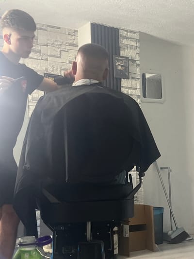 In a dimly lit room, likely a barbershop, a young man with short, styled brown hair stands to the left, focused on cutting the hair of another individual seated in a black barber chair. The seated person, viewed from behind, is wearing a black cape and has a neatly trimmed buzz cut. The barber is holding clippers in his right hand, actively engaged in the haircut. The background features a wall with a grey stone-effect wallpaper and vertical grey panels. A framed picture hangs on the wallpapered section. To the right, a white wall is visible, with a small, recessed mirror-like opening. On the floor, a broom and dustpan stand upright next to a blue trash bin, and a cardboard box is placed nearby. In the foreground, a partially visible green bottle with a purple cap is on a surface. The scene appears to be located in Rudozem, Bulgaria.