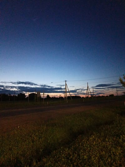 The image depicts a twilight scene at dusk in Kalachevo, Russia. A dark asphalt road occupies the lower third of the frame, bordered by a grassy field. Beyond the road, a line of dark trees forms a silhouette against the horizon. Several wooden utility poles with power lines are visible, some supporting what appear to be road signs. The sky is a gradient of deep blue at the top, transitioning to lighter blues and streaks of orange and pink near the horizon, indicating the sun has recently set. Scattered, dark clouds are present across the lower part of the sky. The overall environment suggests a rural or semi-rural roadside setting. There are no people or animals visible in the image.