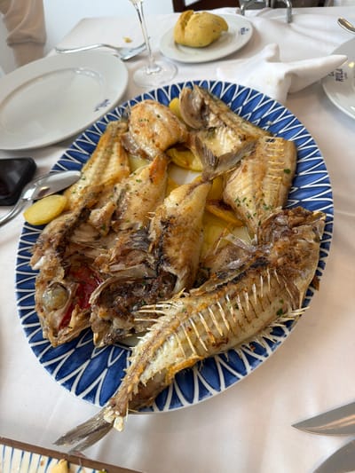 The scene depicts a dining table within a restaurant in Colunga, Spain, identified by "LA RULA LASTRES" branding visible on multiple white plates. A large oval platter, patterned in blue and white, serves as the central object, laden with several whole, cooked fish. These fish possess a golden-brown exterior, with some exhibiting partial consumption, revealing exposed skeletal structures including spines, rib bones, and internal organs. Lemon slices and chopped green herbs garnish the fish, which rest upon visible potato slices. The table is covered with a white fabric tablecloth. Surrounding the main platter are an empty white oval plate bearing a blue logo, a smaller white plate holding a bread roll, a partially visible white plate with the "LA RULA" logo, a clear stemmed wine glass containing liquid, a silver spoon, a silver fork, and a silver knife. A folded white cloth napkin is also present, alongside a partial view of a dark-colored smartphone on the table's edge. No individuals are visible.