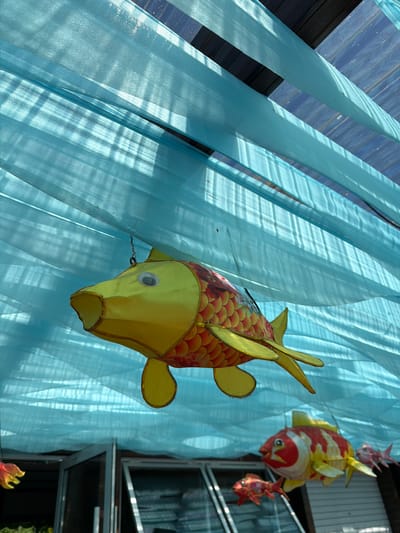 The image captures a vibrant decorative display, primarily featuring fish-shaped lanterns and flowing light blue fabric. In the foreground, a large, yellow and red koi fish lantern is prominently displayed. It has a yellow head with a distinct googly eye, yellow fins, and a body adorned with intricate red and orange scale patterns. The lantern appears to be made of a paper or fabric material, possibly illuminated from within or by ambient light.

In the background, two smaller fish lanterns are visible. One is red and white, positioned above and behind the large yellow fish, while another smaller, reddish lantern is further to the right. These lanterns are suspended against a backdrop of long, translucent strips of light blue fabric, resembling sheer curtains or streamers. The fabric is arranged vertically, creating soft folds and a sense of movement, like water. Strong sunlight casts distinct shadows onto the fabric, suggesting a bright day.

The overall scene evokes a festive and aquatic theme, likely part of a cultural celebration or event in Jiejinkou, China. The koi fish symbolize good fortune and prosperity in East Asian cultures. The light blue fabric reinforces the impression of being underwater or beside a body of water. The photograph is taken from a low angle, looking upwards towards the decorations, with glimpses of dark architectural elements, possibly window frames, in the far background. There is no visible text in the image.