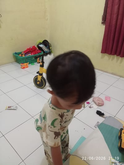 A young child, seen from behind, stands in a room with pale yellow walls and a tiled floor. The child is wearing a light-colored pajama set with a pattern of green and brown animals. In the background, a green plastic basket is filled with toys, including a red toy gun. A yellow and black balance bike with three wheels is positioned on the floor, and a toy train is nearby. To the right, a pink pencil case, keys, and a small pink card are scattered on the floor. A portion of a bed or sofa with a blue and green striped cover is visible in the lower right corner. A window with a dark red curtain is partially visible on the right wall. The timestamp "22/06/2026 21:19" is displayed in the lower right corner of the image. The lighting suggests it is likely evening. The scene appears to be indoors, possibly a child's bedroom or play area in a home.