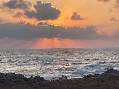 The image depicts a serene coastal sunset scene in the Mate Asher Regional Council, Israel. The sky is a canvas of warm oranges and soft pinks, with scattered gray clouds partially obscuring the setting sun. Rays of sunlight pierce through the clouds, creating a dramatic effect over the water. The sea is calm, with gentle waves lapping at the rocky shoreline. In the foreground, a rocky outcrop borders a patch of dry earth. Two figures, appearing to be a couple, are seated on the rocks, silhouetted against the ocean and the twilight sky, suggesting a moment of quiet companionship. The overall atmosphere is tranquil and picturesque.