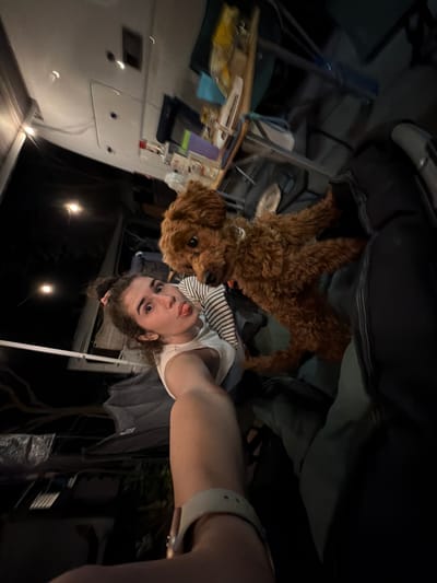 A young woman and a small, brown poodle-type dog are inside a dimly lit campervan. The woman is taking a selfie, sticking her tongue out playfully, with the dog positioned behind her, looking at the camera. The campervan's interior is visible, with various items scattered around, including bottles on a shelf. The scene suggests an informal and relaxed setting, possibly during a camping trip. The image does not provide any information that would indicate a specific location within Πόρτες, Greece.