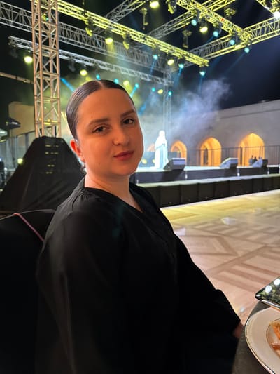 The image features a woman in the foreground, positioned on the right side of the frame, captured from the chest up. She is looking directly at the camera with a neutral expression. She has light skin and dark hair that is neatly pulled back from her face. She is wearing a dark, likely black, garment with long sleeves, which appears to have delicate lace or embroidered detailing along the shoulder or neckline. A faint pink strap or band is visible near her left elbow.
The background reveals a vibrant and dramatic event setting. To the left and behind the woman, there is a large, intricate structure made of metal trusses, characteristic of stage lighting setups. Numerous spotlights are attached to these trusses, emitting bright yellow and blue-green light, creating visible beams through a hazy or smoky atmosphere. Further back and centrally located, there is a large architectural element resembling a building facade with several arched openings, which are warmly illuminated from within. A blurred, light-colored figure, possibly a statue or a person on stage, stands in front of this facade. The stage floor in the upper background shows a light-colored, geometric pattern.
In the upper right corner, partially visible, is a section of a table setting, including a white plate with food and what appears to be silverware, suggesting the woman is seated at a formal dinner or event.
The scene appears to be taking place at night or in a low-light indoor venue, given the prominent stage lighting and dark ambiance. The overall impression is that of an attendee at a large-scale event, possibly a performance, concert, or gala, with a lively and dramatic atmosphere created by the lighting design.