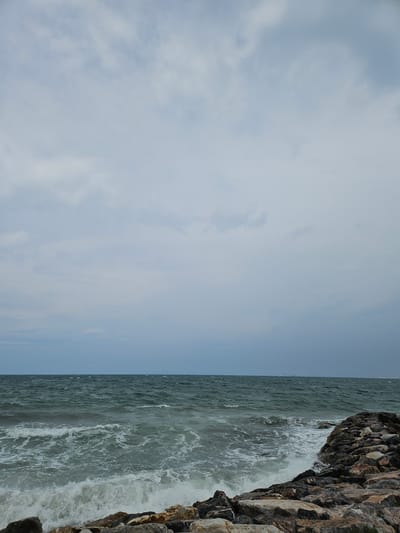 The image shows a rocky shoreline with waves crashing onto the rocks. The sea is choppy and a cloudy sky looms overhead. The scene is likely a coastal area in Güzelbahçe, Turkey, characterized by its natural beauty and the presence of the Aegean Sea. The image is a landscape photograph with no people or animals visible. The focus is on the natural elements of the sea, sky, and rocks.