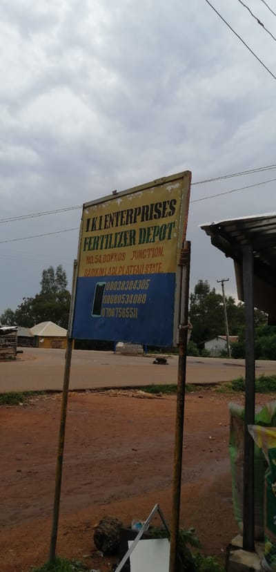 A weathered yellow and blue sign for "LKI ENTERPRISES FERTILIZER DEPOT" stands on two metal poles in front of a dirt road in Kassa Market, Nigeria. The sign has black lettering that lists the address as "NO. 54, BOKKOS JUNCTION, BARIKIN-LADI, PLATEAU STATE." Below the address are three phone numbers: "08038384305," "08080534080," and "07067565511." A stylized image of a smartphone is next to the phone numbers. In the background, there are some trees and buildings, including a structure with a corrugated metal roof. A dark, four-legged animal is lying on the dirt road to the right of the sign. Bags of fertilizer are visible on the far right of the image. The sky is overcast.