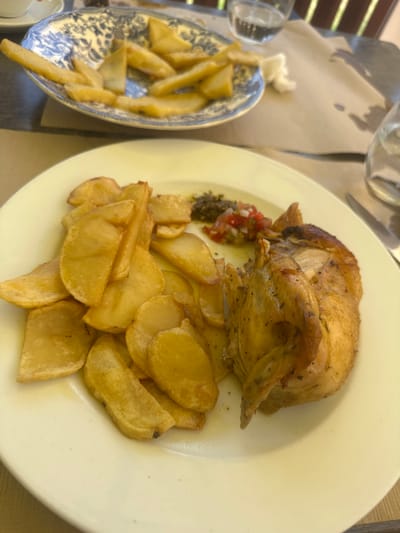 The image shows a close-up of a meal on a white plate, likely at a restaurant in Santa Cristina d'Aro, Spain. The meal consists of roasted chicken, wedges of fried potatoes, and a small portion of chopped salad. The chicken appears to be seasoned with herbs and spices, and is golden brown. The potato wedges are also golden brown and slightly oily. In the background, a blue and white patterned dish filled with more potato wedges is visible, along with two glasses of water and a piece of crumpled white paper. The table setting suggests an outdoor dining experience, possibly on a patio or terrace, given the presence of sunlight and what appears to be a wooden railing in the upper right corner. The focus is on the food, which is presented in a casual, yet appetizing manner.