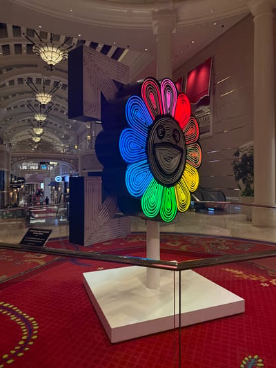 A large, brightly illuminated art installation, approximately 2 meters tall, depicting a stylized flower with a smiling face, is centrally positioned in a grand indoor environment. The flower's petals are formed by concentric neon-like tubes, segmented into distinct colors: red, pink, dark blue, light blue, green, and yellow, radiating from a central face composed of black and white concentric lines. The artwork rests on a white rectangular pedestal, approximately 0.5 meters high, and is contained within a low glass barrier with a dark metallic top rail. The scene is set within a multi-story atrium in Winchester, United States of America, featuring a high, vaulted ceiling adorned with multiple ornate, multi-tiered chandeliers with opaque white shades. Archways and upper-level balconies with decorative metal railings are visible in the background, along with storefronts on lower levels and several indistinct human figures in the distance. Behind the main art piece, a large, white, angular structure with horizontal lines is partially visible. The floor is covered by a red carpet with intricate gold and yellow floral patterns. To the right of the art piece, a light-colored wall with horizontal detailing, a red fabric awning, an escalator, and a potted plant are discernible. A black rectangular sign with white text, partially visible behind the glass barrier, displays the words "MURAKAMI AND FLOWER" and "2019". The overall lighting is bright, combining ambient illumination from overhead fixtures with the internal glow of the art piece.