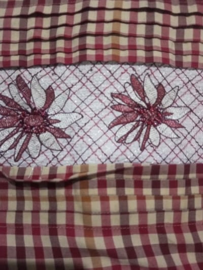 A close-up shows a piece of fabric with alternating red and beige squares. A band of white fabric with an open, square weave is adorned with two floral designs in burgundy and white. The fabric with the squares extends above and below the floral band. The entire piece has a handmade or crafted appearance.