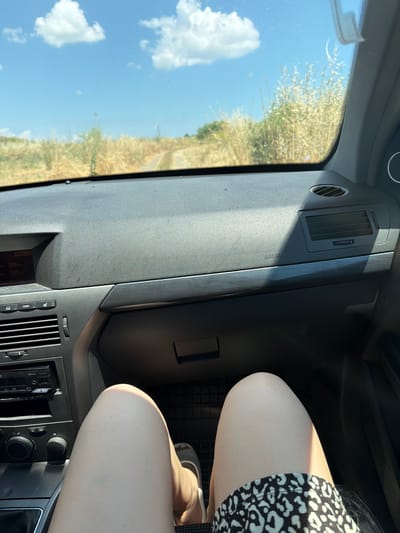 An interior view from the passenger seat of a car looking forward through the windshield. The dashboard of a black car is visible, including the steering wheel, radio, air vents, and an airbag warning. Outside the car, a dirt road winds through dry, grassy terrain under a bright blue sky with scattered white clouds. The sun casts a shadow on the dashboard. A person's legs, clad in a patterned skirt, are visible in the foreground. The car appears to be stopped on the side of a rural road. This scene is consistent with the surroundings of Aheloy, Bulgaria, a town known for its agricultural land and rural landscapes.