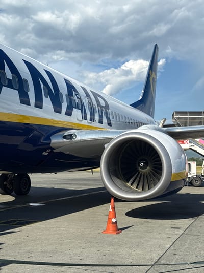 A close-up, low-angle shot captures the engine and fuselage of a Ryanair airplane parked on a tarmac under a partly cloudy sky. The airplane's livery features large navy blue lettering that spells out "RYANAIR" against a white background, with a distinct yellow stripe running along the fuselage. The massive jet engine, made of polished metal, dominates the foreground, its fan blades visible within the intake. A single orange traffic cone with white markings stands in the foreground, casting a shadow. In the background, a portion of an airport jet bridge or staircase and a white service vehicle are visible. The scene is set on a concrete surface marked with yellow lines and other subtle pavement details. The city of Kruja, Albania, is not discernible from the image content, nor are any people or specific actions depicted.