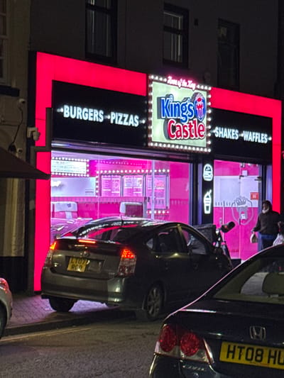 This image depicts a nighttime street scene in Preston, United Kingdom, specifically focusing on the brightly lit exterior of "King's Castle," a fast-food establishment located at 30-31 Church Street. The building's dark facade is prominently outlined by vivid pink-red neon strip lighting. The central illuminated sign displays "King's Castle" in blue and red lettering, featuring a castle logo and surrounded by individual bulbs, with the tagline "Home of the King" inscribed above. Adjacent black panels, also outlined in pink-red neon, advertise "BURGERS * PIZZAS" on the left and "SHAKES * WAFFLES" on the right in white text. The interior, visible through large glass panels, is saturated with pink-magenta lighting, revealing various menu displays and white seating arrangements. A white security camera is mounted on the wall to the left of the storefront.
Three vehicles are present on the street. A dark grey Toyota Prius (UK registration LY07 RCQ, issued between March and August 2007) is parked or stopped in the middle ground, its rear facing the viewer with illuminated taillights. In the immediate foreground on the right, the rear section of a dark blue/black Honda Civic (UK registration HT08 HU, issued between March and August 2008) is visible, also with illuminated taillights. A small portion of a light-colored vehicle's front bumper is discernible on the far left.
One individual is observed exiting or standing near the right entrance of King's Castle, facing left. The person is wearing a dark jacket, dark trousers, and a dark face covering, and carries a light-colored takeout package or box in their right hand. A dark, tall, upright object, possibly a delivery stand or sign, is positioned on the pavement near the right-hand Honda Civic. Pavement and building details are visible under and around the vehicles and storefront.
