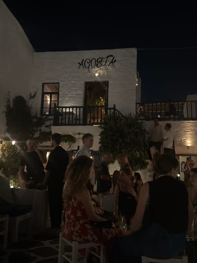 The image shows an outdoor scene at night, likely a restaurant or bar area in Naousa, Greece. Several people are visible, mostly gathered around tables. The architecture suggests a whitewashed building with dark window frames and balconies, typical of Greek island style. A sign with the word "ACOSTA" is prominently displayed in black lettering on the white wall.

The atmosphere appears to be social, with people engaged in conversation and possibly dining or drinking. Some individuals are standing, while others are seated. The lighting is dim, with some artificial lights illuminating the area and casting glows from the windows, suggesting it's evening or night. The presence of plants and trees adds to the outdoor setting. The overall impression is of a lively, social gathering at a restaurant on a warm night.