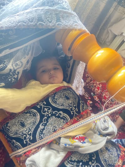 An infant is positioned supine within a traditional, possibly wooden, cradle or *beshik*, veiled by a light-colored, lace-trimmed netting. The baby wears a solid light-yellow long-sleeved garment and rests upon a dark blue fabric with silver-gold ornate patterns. To the right of the infant, a prominent, multi-segmented, turned wooden object, finished in a golden-yellow hue, is visible; this is consistent with a *beshik* component. The background features various patterned textiles in red and dark tones, suggesting an indoor domestic environment. A thin, light-colored braided strap with blue accents extends across the lower right frame over additional textile items. The infant gazes directly forward with a neutral expression. The scene is located in Galaosiyo, Uzbekistan.