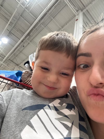 A close-up selfie captures a young boy and an adult woman in a shopping cart. The boy, on the left, has light brown hair and is wearing a gray t-shirt with a Nike logo. He is looking downwards with a slight smile. The woman, on the right, is also looking towards the camera with her lips puckered and one eye squinted. The background shows the ceiling of a large store with industrial lighting and exposed pipes. Several items are visible in the shopping cart behind the boy, including a blue bag and a loaf of bread with striped packaging. The setting appears to be an indoor retail environment, possibly a supermarket or warehouse store. The image was taken in the City of Yonkers, United States of America.