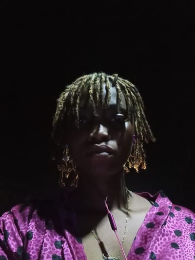 A woman with blonde dreadlocks is shown in the image, wearing a pink patterned top. She is also wearing a gold chain necklace and has a pink object on her chest. The image appears to be taken in a low-light environment. The location in the city of Ogbe, Nigeria is not discernible from the image itself.
