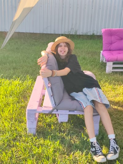 A young person is lounging on a pink-painted pallet chair outdoors in the grass. They are wearing a straw hat, a black t-shirt, light blue shorts with red and white stripes, and black high-top sneakers with white laces and white ruffled socks. The person is sticking their tongue out playfully at the camera, with their arms resting on the chair and bracelets on one wrist.
In the background, there is a white corrugated metal fence, a partially visible light-colored tarp or tent, and another pink pallet chair with a cushion. The scene is set in a grassy area, likely a yard, with the sun casting long shadows, suggesting it is late afternoon or early evening. The lighting is warm and the grass appears green and lush, indicating a pleasant weather day. The location is Nechaiane, Ukraine. There is no visible text in the image.
