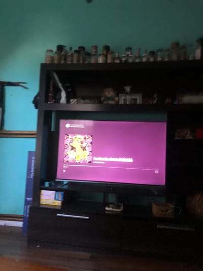 The image shows an indoor scene in what appears to be a living room. A dark-colored entertainment center dominates the frame, housing a television displaying a purple screen with abstract artwork and text. The entertainment center contains various objects on its shelves, including decorative bottles, books, and other miscellaneous items. In the background, a teal-colored wall is visible. Additionally, parts of a picture frame can be seen in the lower part of the image.
The location is identified as Beccar, Argentina. However, there are no specific details in the image to confirm this.
No people, actions, or interactions are visible in the image.