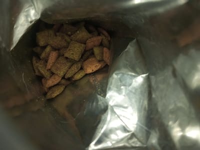 The image shows a close-up, interior view of a shiny, metallic bag, possibly made of foil. At the bottom, a pile of small, irregularly shaped pieces, likely food, are visible. These pieces are primarily green and brown, suggesting a mix of ingredients. The lighting creates strong highlights and shadows on the bag's surface, emphasizing its crumpled texture. The overall composition focuses on the textures and colors within the bag's limited space.