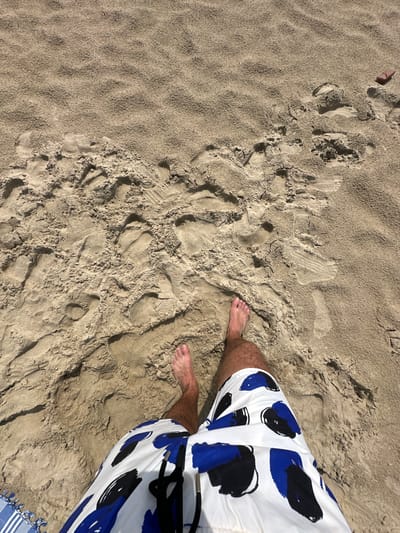 The image shows a top-down view of a sandy beach. The sand is light brown and textured with numerous footprints. In the foreground, a person's legs and feet are visible. They are wearing white swim trunks with a blue and black abstract pattern. The person's bare feet are resting on the sand. In the upper right corner of the image, a small brown object, possibly a piece of driftwood or a shell, is visible on the sand. In the lower left corner, a portion of a blue and white striped towel can be seen. The lighting suggests a bright, sunny day. This scene is consistent with a location like the beach in Koksijde, Belgium, which is known for its sandy shores.
