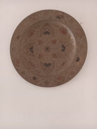 A tarnished, circular, decorative plate with intricate floral and geometric patterns is positioned against a plain, off-white background. The plate exhibits a color palette of subdued browns, reds, and blacks. The surface is aged and textured. The location is unknown and there is no evidence to support this image being taken in the city of Ksar-el-Kebir, Morocco.