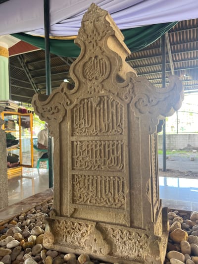 A weathered, stone gravestone with intricate Arabic calligraphy is centrally positioned within a grave enclosure in Beuringen, Indonesia. The gravestone, which appears to be ancient, is adorned with decorative carvings and rests on a bed of smooth, light-colored river stones. Behind the gravestone, a protective canopy is supported by dark metal poles and draped with white and green fabric. In the background, a bookshelf filled with books is visible through a glass pane, and beyond that, a person in a light-colored shirt is seated on a green chair. The environment suggests a burial site or a historical marker, possibly within a mosque complex or cemetery, with the background indicating a semi-outdoor or sheltered area.