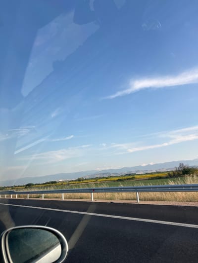 The image depicts a view from a vehicle traveling on a highway near Saraia, Bulgaria. The foreground shows the asphalt road with a white lane marker, and a side-view mirror of the vehicle. A metal guardrail separates the road from a field of dry grass and a distant field of yellow flowers, likely sunflowers. In the background, a range of mountains is visible under a bright blue sky with scattered wispy clouds. The scene is illuminated by daylight, suggesting it was taken during the day. There are no people or animals visible in the frame.