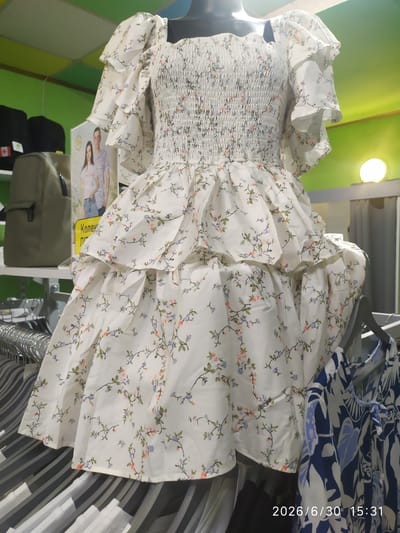 This media file shows a white dress with a floral pattern of small blue and pink flowers and green vines. The dress has a smocked bodice and puffy sleeves. It appears to be displayed on a mannequin in a retail setting. To the right of the dress, another garment with a blue and white abstract floral pattern is visible, hanging on a rack.
In the background, there is a bright green wall and a light fixture. Below the dress, a postcard-sized advertisement is visible, featuring a man and a woman. Some text is visible on the advertisement, including "Конек". The overall scene suggests an indoor environment, likely a clothing store.
The timestamp "2026/6/30 15:31" is overlaid on the right side of the image. The scene is brightly lit, suggesting it is daytime.