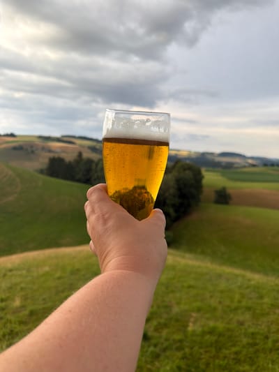 The image shows a hand holding a glass of beer against a backdrop of rolling green hills and a cloudy sky. The beer is golden yellow and has a foamy head. The background depicts a rural landscape with fields, trees, and rolling hills, indicative of a natural or agricultural setting. The sky is overcast with some bright patches, suggesting either early morning or late afternoon light. The scene evokes a sense of relaxation and enjoyment, possibly a person savoring a drink outdoors in the countryside. There is no visible text in the image. The location context of Ufhusen, Switzerland, further suggests a rural European landscape.