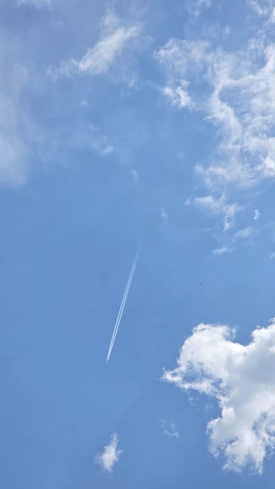 The image presents a wide-angle view of a bright, clear blue sky, partially adorned with white clouds. The sky dominates the frame, showcasing varying shades of blue indicative of a sunny daytime.
Scattered across the expanse are numerous white clouds. In the upper left quadrant, there are wispy, diffused cirrus clouds, while the upper right features a more prominent, distinct cumulus cloud formation that appears fluffy and well-defined. Smaller, dispersed cloud patches are also visible throughout the frame.
A notable element is the long, linear white contrail streaking diagonally across the sky from the lower-left to the upper-right. This contrail signifies the passage of a high-flying airplane, which itself is not clearly visible beyond the trail. Slightly above and to the left of the contrail's mid-section, a very small, dark, indistinct speck can be observed, possibly a bird or another distant object.
The scene evokes a feeling of openness and tranquility, suggesting a pleasant, fair-weather day. The absence of direct sunlight sources or distinct shadows implies it could be late morning or early afternoon. No visible text is present in the image. The setting for this serene sky view is Drenkovo, Bulgaria.