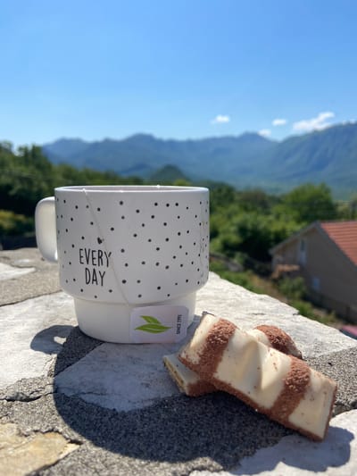 A white mug with black polka dots and the words "EVERY DAY" sits on a stone surface, accompanied by two Kinder Bueno bars. The mug has a tea bag with a green leaf logo and the text "SINCE 1991" attached to it. The backdrop includes a view of green trees, mountains, and a clear blue sky, possibly a scene within the city of Sotonići, Montenegro. A house with a red tile roof is partially visible below the trees.